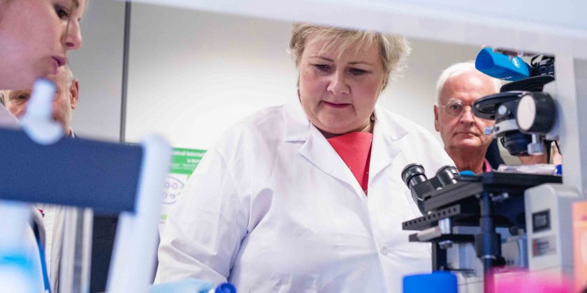 Woman in lab coat behind microscope sourounded by people