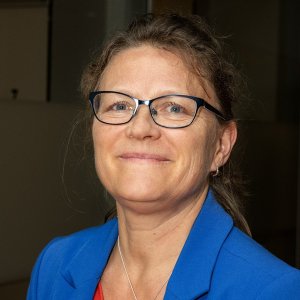 Åslaug Helland is looking into the camera with a content smile, wearing a blue jacket and glasses. 