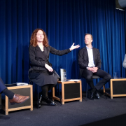 Three people in a discussion on a stage, sitting, one of which is Camilla Stoltenberg, who is engaged with her arm in a gesture.