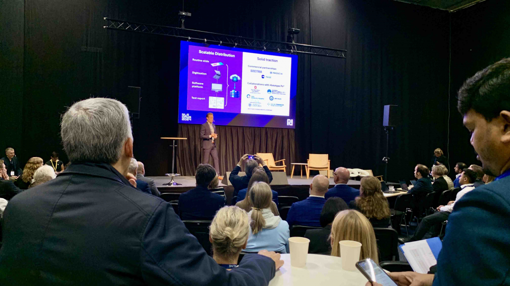 Man on a stage in front of screen and audinece. 